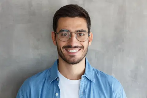 Smiling man with dark hair, beard, and glasses in a denim shirt; TN Capital Investments in Naples, FL, gray backdrop.