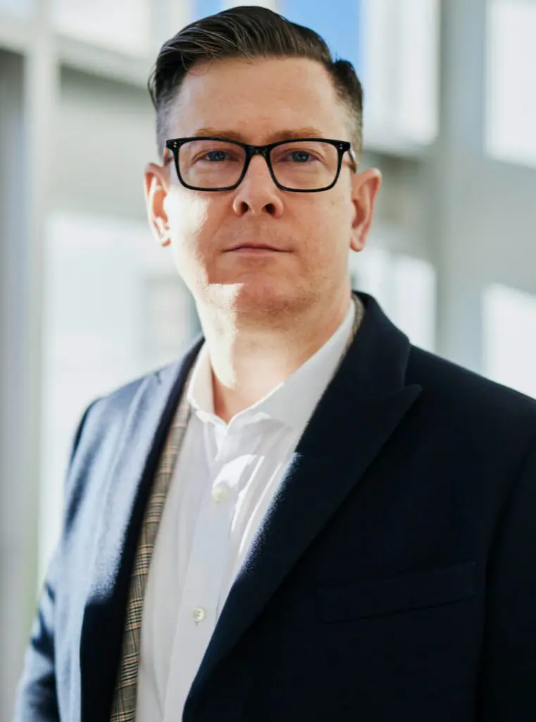 Thomas Portmann of TN Capital Investments in Naples, FL, wears glasses, a white shirt, and dark blazer indoors by windows.