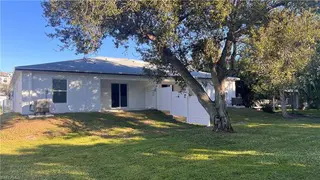 Single-story white house with gray roof and blue door in Naples, FL, listed by TN Capital Investments; fenced yard and large tree.