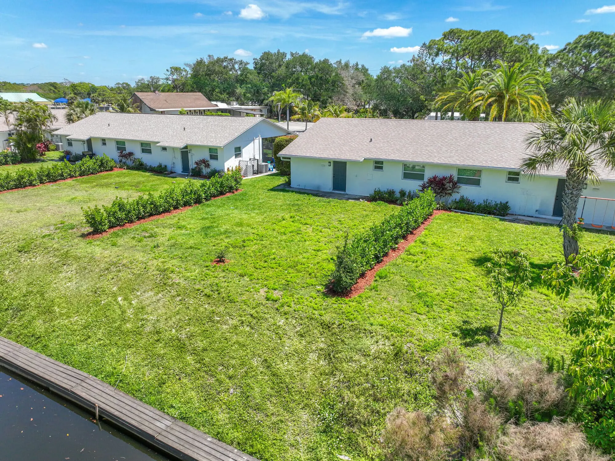 Two white buildings with gray roofs, managed by TN Capital Investments in Naples, FL, are divided by a lawn near a canal.