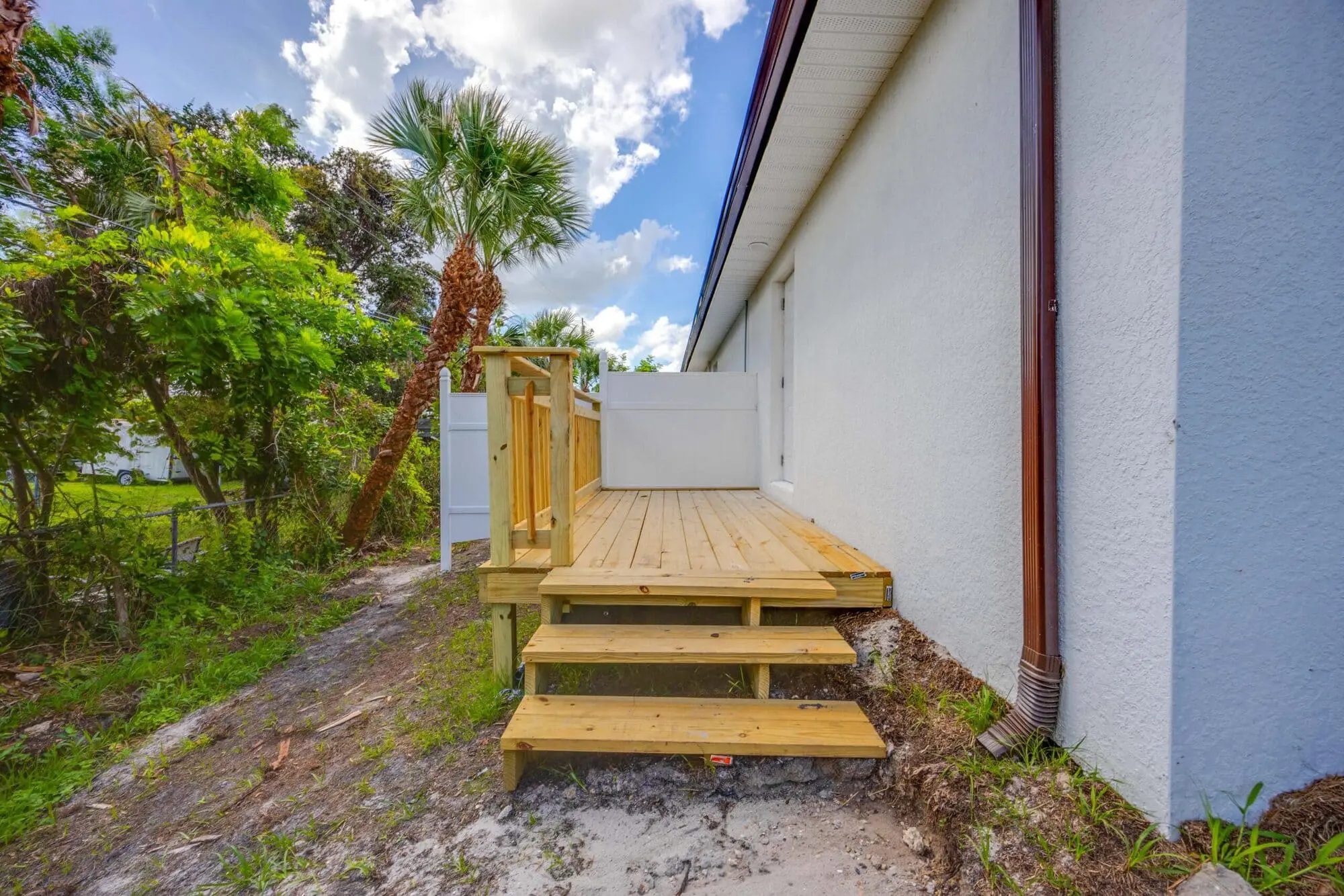 A small wooden deck with three steps by a white building, greenery, and sandy ground at a TN Capital Investments site in Naples, FL.