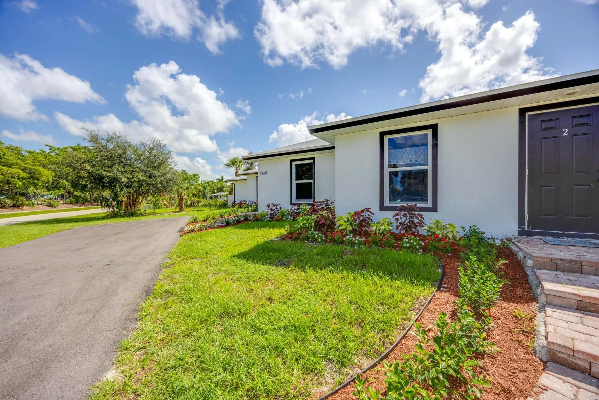 A single-story white house with black trim by TN Capital Investments in Naples, FL, features a black door and green landscaped yard.