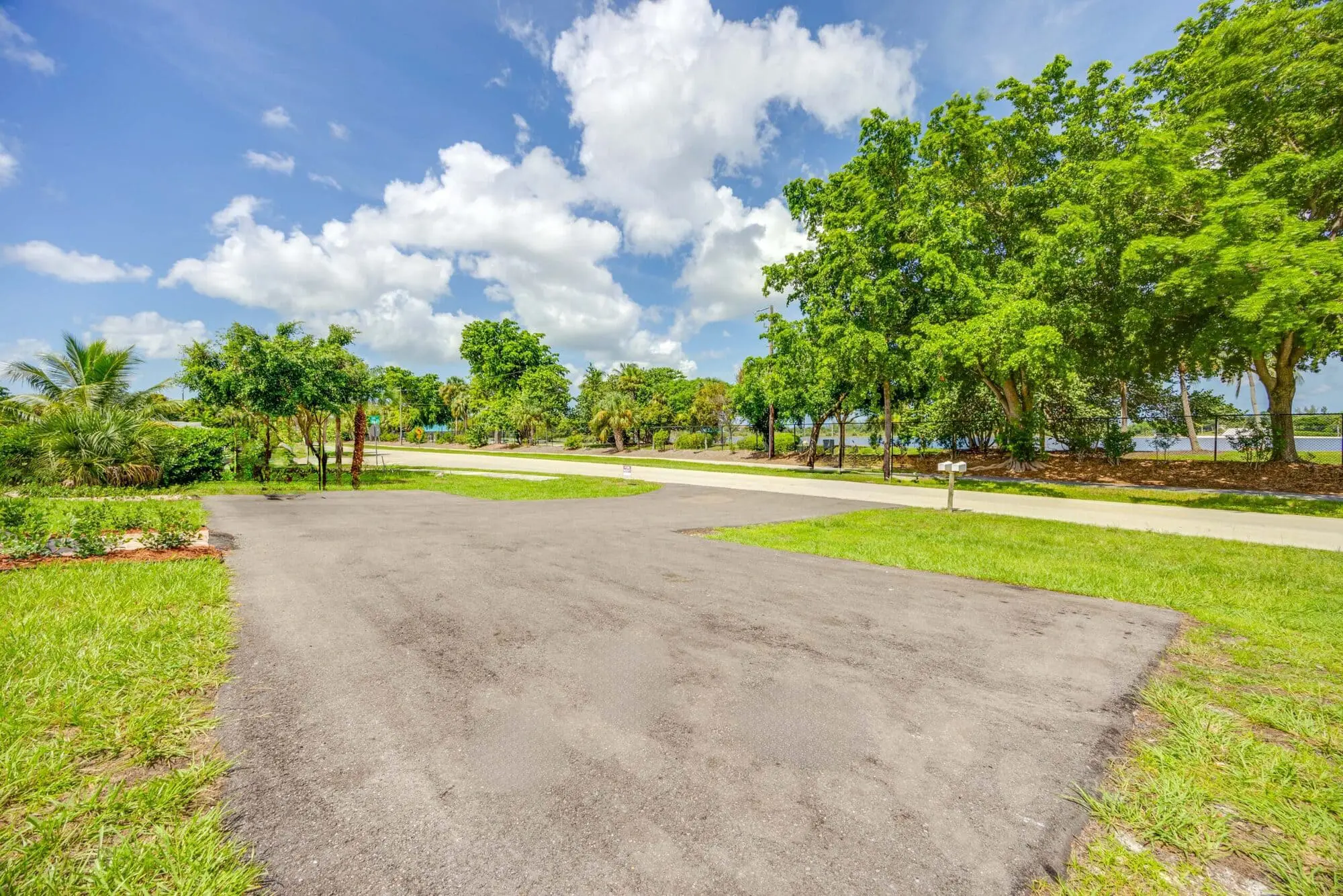 A paved driveway with green grass and trees, a road, and partly cloudy sky is shown by TN Capital Investments in Naples, FL.