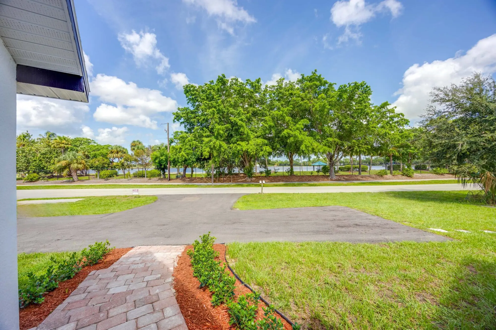 Paved walkway, driveway, green lawn, and water view through trees under a partly cloudy sky for TN Capital Investments in Naples, FL.